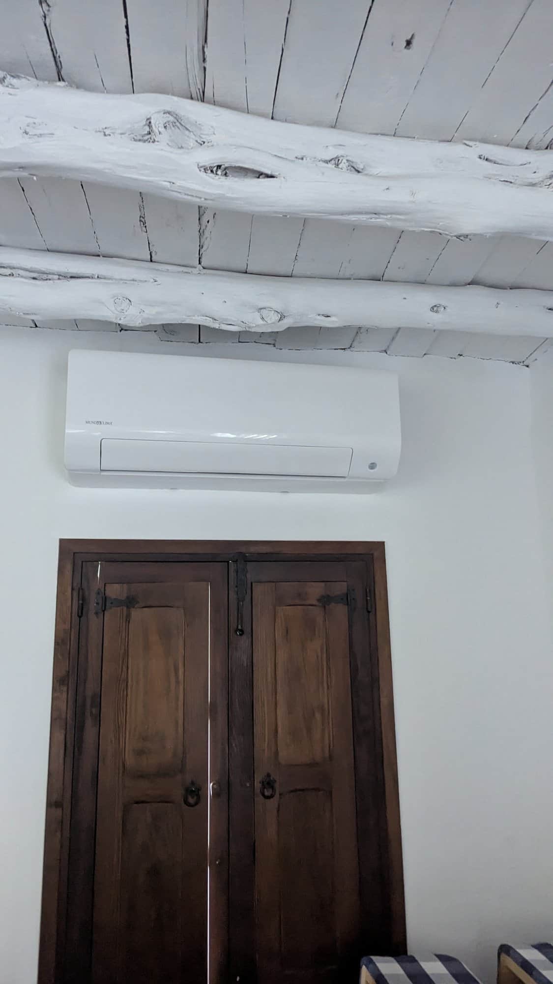 Airco-wandunit boven een houten deur, onder een wit balkenplafond.