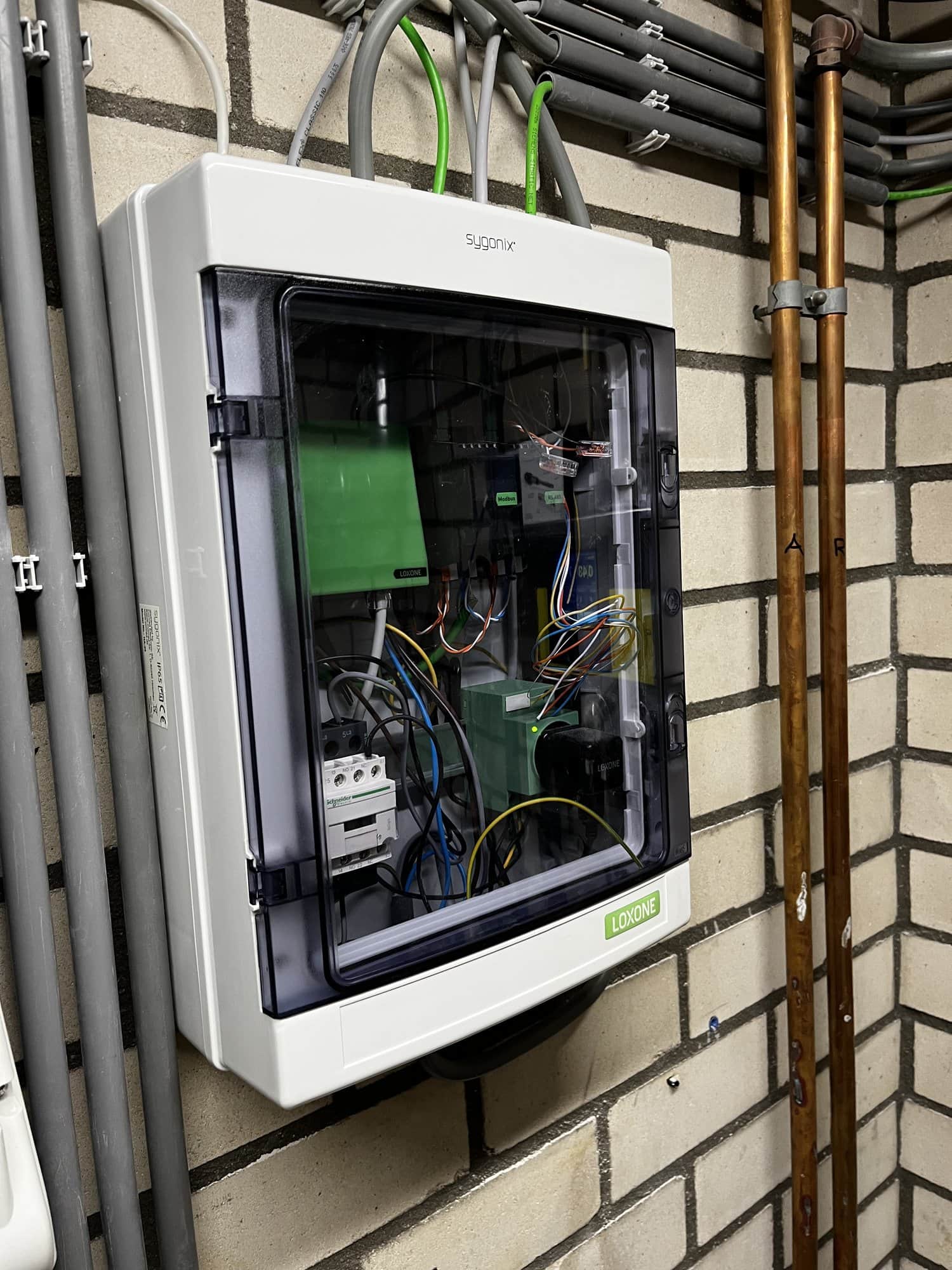 Opbouwkast met Loxone-componenten en invoerende bekabeling op een gemetselde wand.